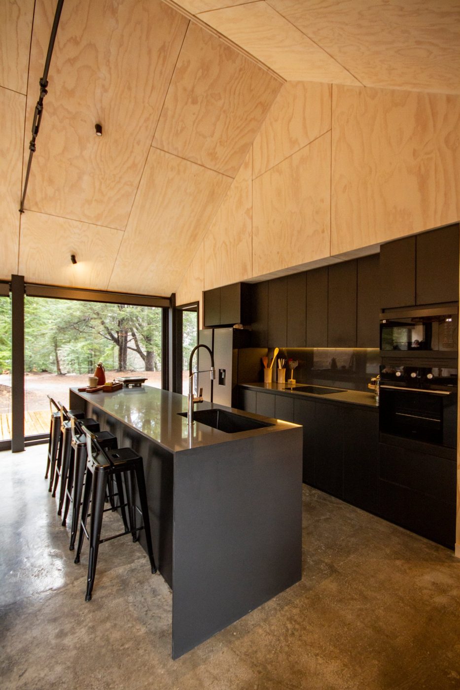 Cocina negra con isla y cielo de madera de la casa Malalcahuello, con ventanal hacia el bosque